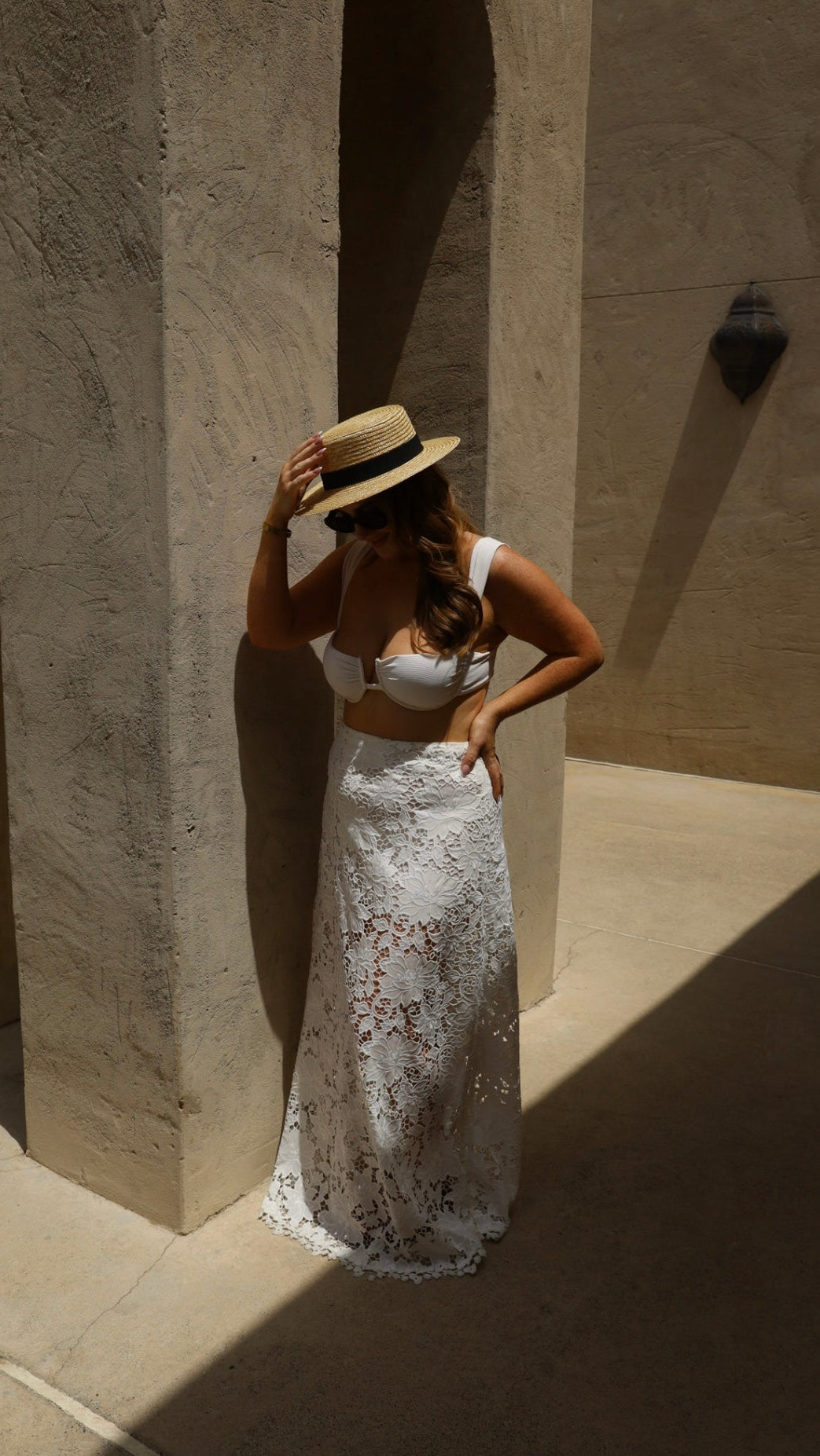 Woman in a white lace dress and straw hat standing against a stone wall.