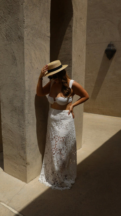 Woman in a white lace dress and straw hat standing against a stone wall.